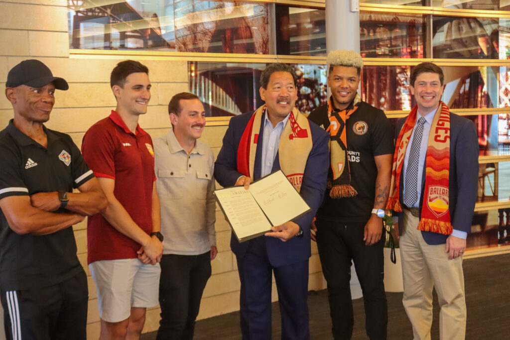 Mayor Bruce Harrell, Councilmember Dan Strauss, and members of the Ballard FC organization pose for photos with the proclamation. 