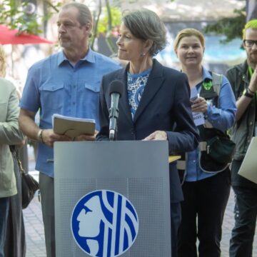 Council President Sara Nelson speaking at a podium in Occidental Square Park, surrounded by clapping supporters of her new legislations