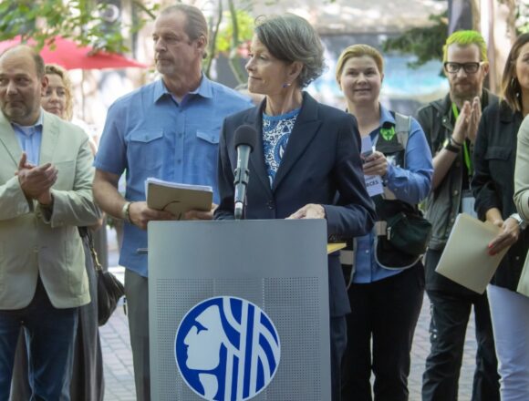 Council President Sara Nelson speaking at a podium in Occidental Square Park, surrounded by clapping supporters of her new legislations