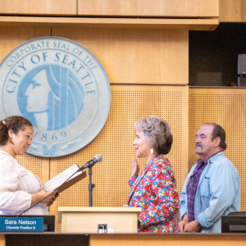 Debora Juarez raising her right hand in front of microphone while the Seattle City Clerk swears her in and a man stands next to her
