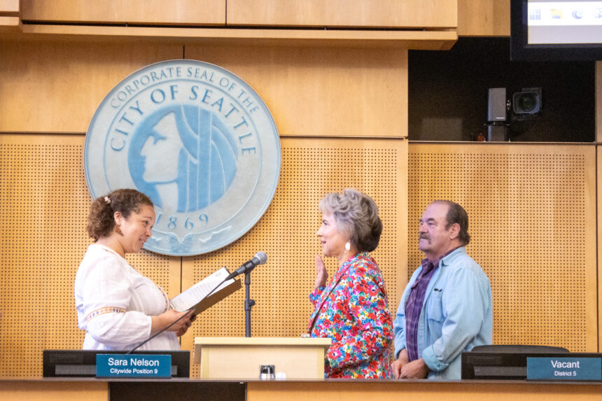 Debora Juarez raising her right hand in front of microphone while the Seattle City Clerk swears her in and a man stands next to her