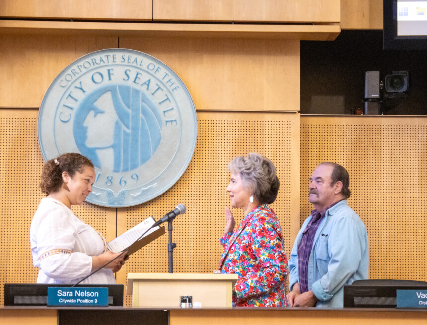 Debora Juarez raising her right hand in front of microphone while the Seattle City Clerk swears her in and a man stands next to her