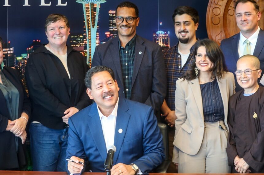 Mayor Harrell sitting, smiling and holding a pen while surrounded by a group of people standing and smiling. The group includes Councilmember Rinck, Councilmember Saka, and other stakeholders to the bill