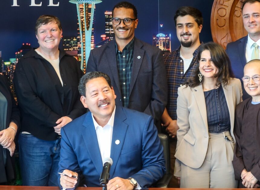 Mayor Harrell sitting, smiling and holding a pen while surrounded by a group of people standing and smiling. The group includes Councilmember Rinck, Councilmember Saka, and other stakeholders to the bill