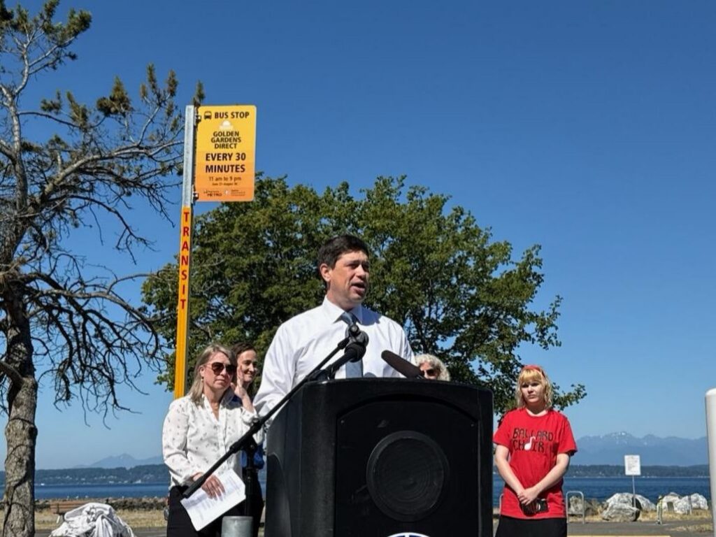 Councilmember Strauss speaking at a podium in front of a bus stop sign for Golden Garden Direct