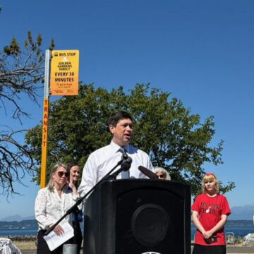 Strauss Speaking at podium in front of a Golden Gardens Direct Bus sign Strauss Speaking at podium in front of a Golden Gardens Direct Bus sign