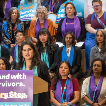 Councilmember Rinck speaking at a podium with a sign that reads "Stand with Survivors. Every Step.", many people in the background wearing orange, purple, and teal