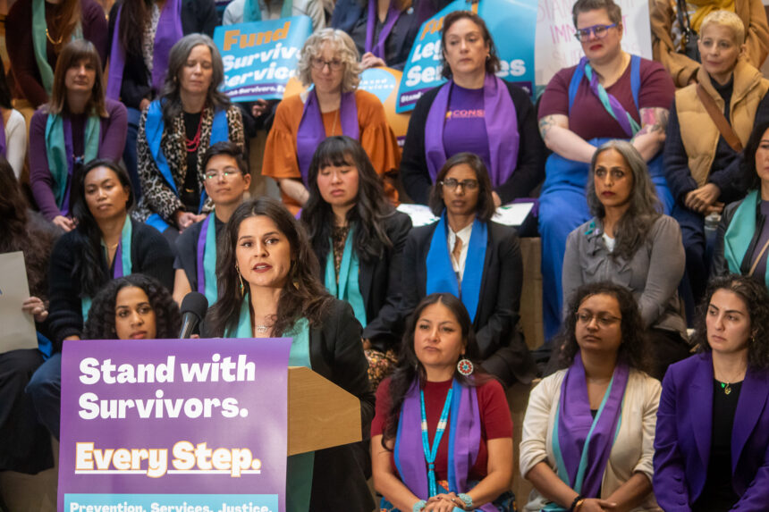 Councilmember Rinck speaking at a podium with a sign that reads "Stand with Survivors. Every Step.", many people in the background wearing orange, purple, and teal