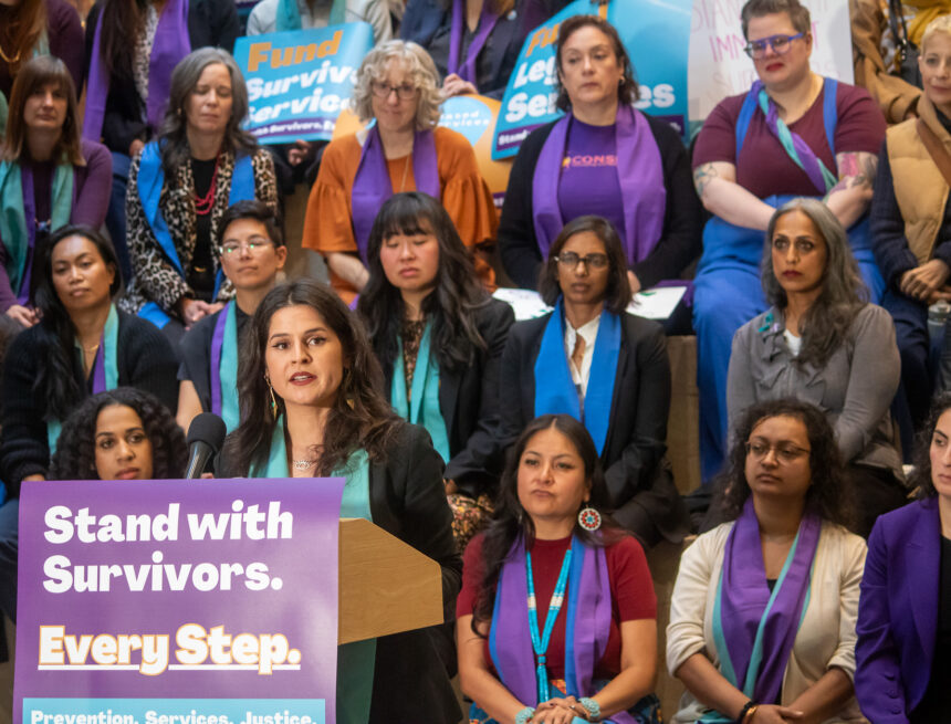 Councilmember Rinck speaking at a podium with a sign that reads "Stand with Survivors. Every Step.", many people in the background wearing orange, purple, and teal